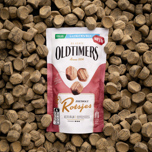 Oldtimers Zoethout Rotsjes suikervrije vegan snoepjes in een staande verpakking, omringd door zoethoutblokjes op een houten ondergrond.